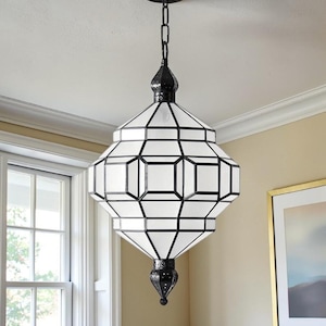 May include: A hanging pendant light fixture featuring a geometric, multi-faceted white glass shade encased in a dark metal frame. The lantern-style lamp is suspended by a black chain and includes decorative metal finials at the top and bottom.