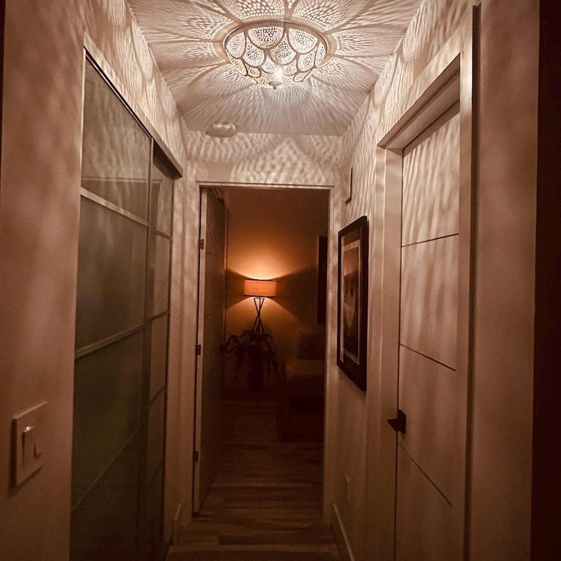 May include: A white semi-flush mount ceiling light fixture with a scalloped, perforated metal shade that casts intricate, petal-shaped shadows across the walls and ceiling of a narrow hallway with light-colored doors and wood-look flooring.