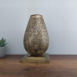 May include: A gold metal table lamp with a decorative, intricate, cut-out design. The lamp has a round, teardrop-shaped base and a square base.