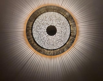 May include: A circular wall sconce featuring a central solid dark disc surrounded by an intricate, perforated metal pattern of swirling floral motifs. The fixture emits a warm, radial light that casts distinct, elongated shadows on the wall.
