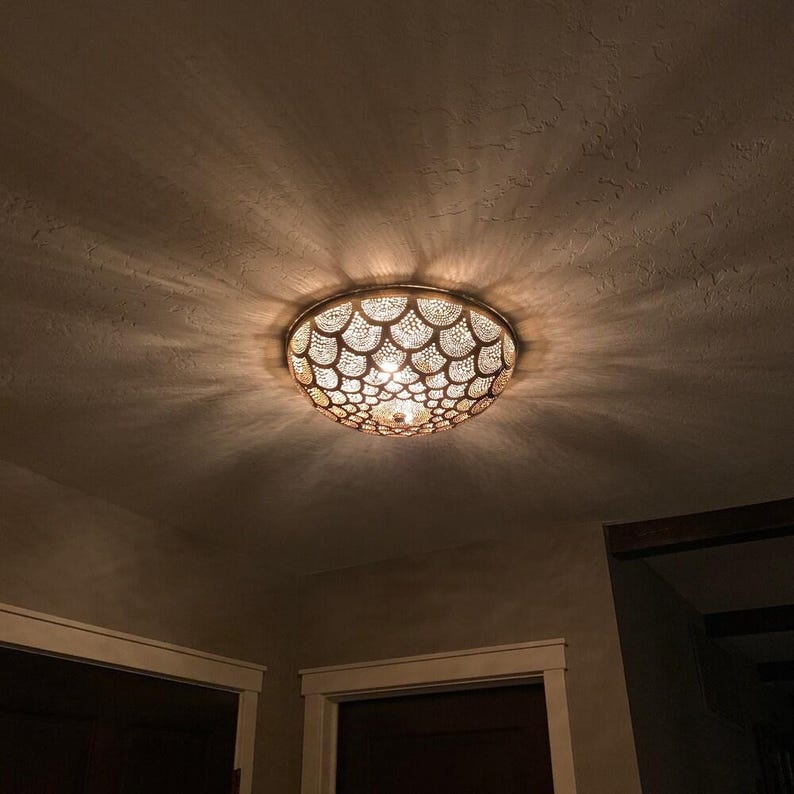 May include: A decorative ceiling light fixture with a bronze finish. The light has a circular shape with a detailed, patterned design. The light is turned on, casting a warm glow and intricate shadows on the ceiling.