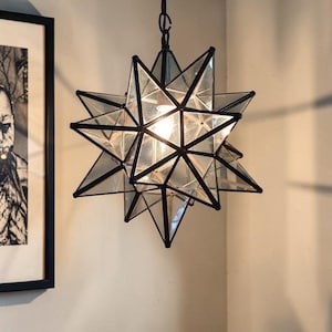 May include: A star-shaped pendant light fixture with a black metal frame and clear glass panels. The light is illuminated, casting a warm glow. The fixture is hanging from a chain. The style is modern and geometric.