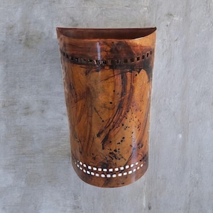 May include: A semi-cylindrical wall sconce featuring a mottled copper and dark brown finish with brushstroke textures. The fixture includes horizontal bands of small, square cutouts along the top and bottom edges, creating a decorative pattern.