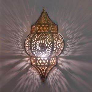 May include: An elaborate gold-toned wall sconce featuring detailed cut-out designs. The lamp's pattern includes geometric and floral elements, creating decorative light shadows on the wall. The sconce has a pointed top and bottom, and a central light source.