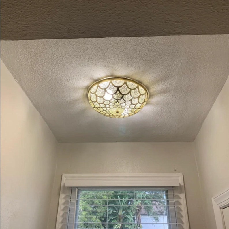 May include: A circular flush mount ceiling light fixture featuring a gold-toned metal frame with an intricate, repeating scalloped pattern. The light emits a warm glow through the cutouts, casting a decorative shadow pattern on the ceiling.