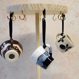 May include: A wooden mug rack with brass hooks, displaying three ceramic mugs. One mug is brown and white striped, another is black and white, and the third is beige with black floral designs. Each mug hangs from a black ribbon.