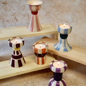 May include: A collection of ceramic candle holders in various colors, including pink, orange, blue, and purple. Each holder has a unique striped design and holds a lit tea light candle. The holders are arranged on wooden shelves.