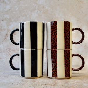 May include: Four ceramic mugs stacked in pairs. Two mugs feature black and white vertical stripes, while the other two have a cream background with brown leopard print stripes. All mugs have black or brown handles.
