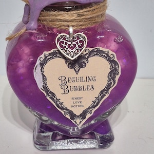 Puede incluir: Una botella de poción morada en forma de corazón con una etiqueta decorativa que dice "Beguiling Bubbles, Finest Love Potion". La botella está sellada con cera morada y cuerda, y un dije de corazón plateado cuelga del cuello.