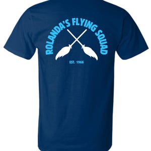 May include: Navy blue t-shirt with a light blue and white graphic on the back. The graphic features crossed brooms and the text "ROLANDA'S FLYING SQUAD" in a semi-circle, with "EST. 1966" below. A casual, crew neck tee.