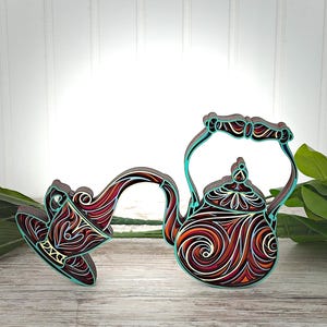 May include: A layered wooden wall art piece depicting a teapot pouring liquid into a teacup. The design features intricate swirling patterns in shades of deep red, orange, white, and black, with a vibrant turquoise outline defining the shapes.