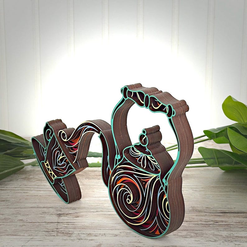 May include: A layered wooden wall art piece shaped like a teapot and a teacup, featuring intricate cutouts with brown, teal, orange, and yellow accents. The design displays a swirling, decorative pattern throughout the structure of the teapot.