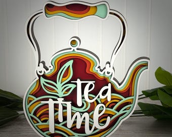 May include: A multi-layered wooden wall art piece shaped like a teapot, featuring a stacked design with colors of mint green, yellow, orange, and deep red. The white text "tea time" is layered over the center, accompanied by a leaf graphic.
