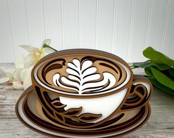 May include: A layered wooden wall art piece shaped like a coffee cup on a saucer. The design features stacked cutouts in shades of brown, tan, and white, creating a three-dimensional effect with a white latte art pattern on the coffee surface.