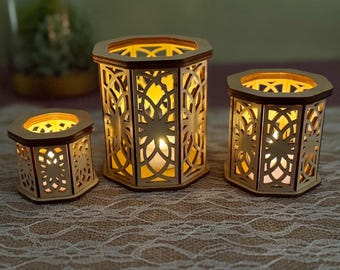 May include: Three wooden candle lanterns with intricate cut-out designs. The lanterns are hexagonal, with a warm glow emanating from within. The smallest lantern is 3 inches tall, the medium is 5 inches, and the largest is 6 inches.