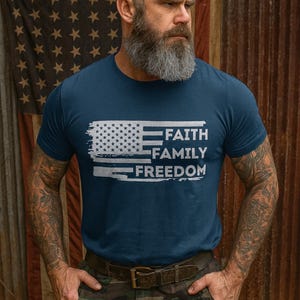 May include: Navy blue t-shirt featuring a distressed American flag design and the words "FAITH FAMILY FREEDOM" in white. The shirt is made of a soft material and is suitable for casual wear. The background features an American flag and a rustic metal wall.