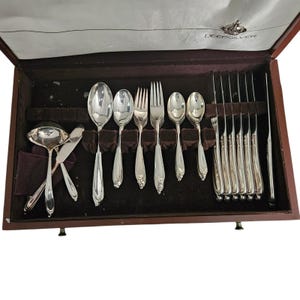 May include: A silver-plated flatware set in a brown wooden storage case with a white satin lining. The set includes a ladle, knives, forks, and spoons, all featuring a decorative handle design. The interior lid displays the text DEEPSILVER.