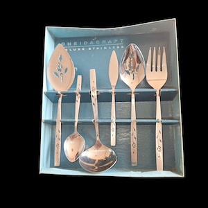May include: A set of stainless steel flatware by OneidaCraft, Deluxe Stainless, in a blue box. The set includes a serving spoon, a slotted spoon, a ladle, a knife, a fork, and two other serving utensils. Each piece has a floral design.