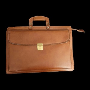 May include: A brown leather briefcase with a handle and a gold-colored clasp. The briefcase has a flap closure and a zipper on the side. The leather has a slightly worn appearance.
