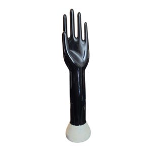 May include: A black, glossy hand sculpture with outstretched fingers. The hand is attached to a long, cylindrical arm that ends in a white, rounded base. The sculpture is likely made of ceramic or a similar material.