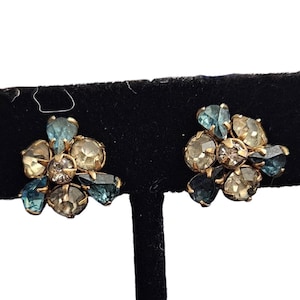 May include: A pair of vintage clip-on earrings. Each earring features a cluster of faceted, teardrop-shaped, and round gemstones in shades of blue, champagne, and clear, set in a gold-tone metal. The earrings are displayed on a black surface.