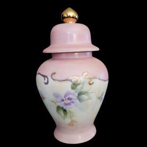 May include: A pink ceramic jar with a lid, featuring hand-painted purple pansies and gold accents. The jar has a gold finial on top and is set against a black background. The floral design adds a touch of elegance.