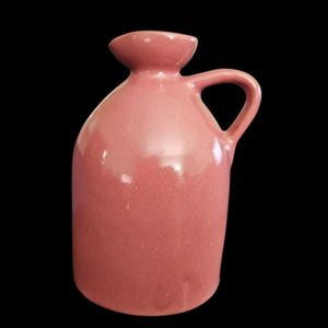 May include: A pink ceramic pitcher with a handle and a flared spout. The pitcher has a rounded body and a smooth, glossy finish. The handle is a simple loop shape, and the spout is slightly wider than the neck.