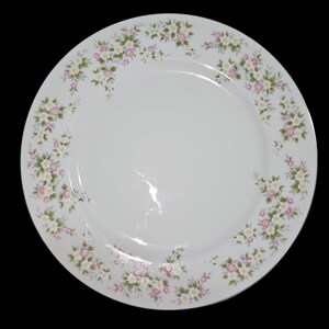 May include: A round white ceramic dinner plate featuring a delicate floral pattern along the rim. The design consists of small clusters of pink and white flowers with green leaves arranged in a repeating motif around the edge of the plate.