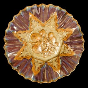 May include: A round ceramic plate with a scalloped edge, featuring a central beige relief design of fruit including pears and grapes. The plate has a glossy glaze with deep purple and blue tones, accented by brown highlights on the star-shaped rim.
