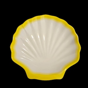 May include: A white, shell-shaped dish with a bright yellow rim. The dish has a scalloped edge and a ribbed interior design. The background is solid black, highlighting the dish's shape and color.