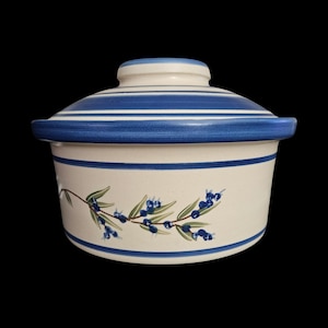 May include: A round ceramic casserole dish with a matching lid, featuring a cream-colored base with horizontal blue stripes. The exterior is decorated with a painted botanical motif of green leaves and small blue berries encircling the center.