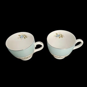 May include: Two vintage teacups with a light blue band and silver trim. Each cup features a floral design with blue and green accents. The cups have a white interior and a curved handle.