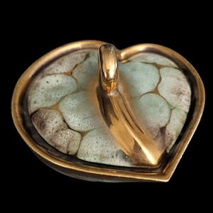 May include: A heart-shaped ceramic dish featuring a crackled, pale green and white glaze pattern. The dish is framed by a smooth, metallic gold-colored rim and includes a prominent, curved gold-colored handle extending from the center.