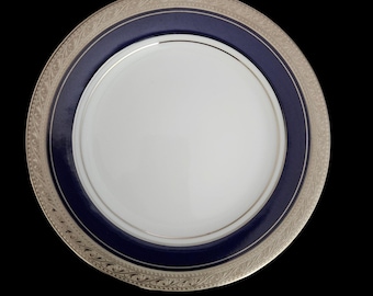 Op de afbeelding: Een rond dinerbord met een effen wit midden, een brede marineblauwe band en een buitenrand versierd met een ingewikkeld zilverkleurig metalen filigraanpatroon. Het bord heeft een gladde, glanzende afwerking en een strakke, ronde vorm.