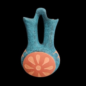 May include: A turquoise ceramic vase with a unique double-spout design and a central opening. The vase features a terracotta-colored circular design with a floral pattern. The vase is set against a black background.