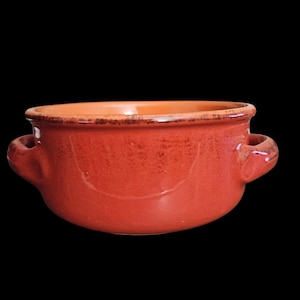May include: A rustic, red ceramic bowl with two small handles. The bowl has a textured surface and a contrasting orange interior. The rim has a darker, aged appearance. This bowl is suitable for serving soups, stews, or other dishes.
