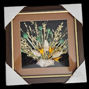 May include: A framed dried flower arrangement. The artwork features a variety of dried flowers in shades of green, yellow, and beige, set against a black background. The frame is dark brown with a gold inner border, and is protected by cardboard corner protectors.