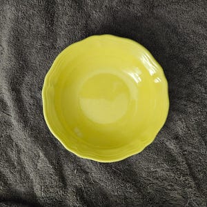 May include: A bright yellow ceramic bowl with a scalloped edge. The bowl has a smooth, glossy finish and is set against a dark gray textured background. The bowl is round and appears to be empty.