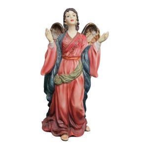 May include: A standing angel figurine with brown hair, wearing a long pink robe with a dark blue textured shawl and a green sash. The figure has large, detailed brown wings, arms raised with open palms, and is looking upward with a serene expression.