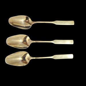 May include: Three silver-toned metal spoons arranged vertically. Each spoon features an oval-shaped bowl and a flat, rectangular handle. The polished surfaces reflect light, showing a smooth, metallic finish on all three utensils.