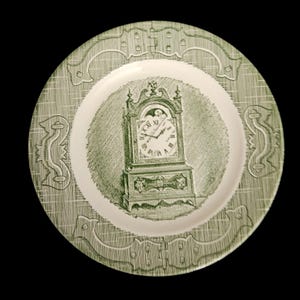 May include: Green and white decorative plate featuring a detailed illustration of a grandfather clock. The plate has a textured border with decorative scrollwork and a white center.