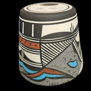 May include: A cylindrical ceramic vessel with a black rim and base, featuring a geometric design in black, white, gray, orange, and blue. The artwork includes stripes, triangles, and curved lines, with a textured gray base.