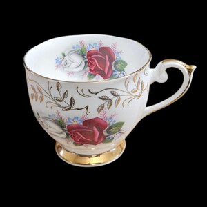 May include: White teacup with a gold rim and handle, featuring a floral design with red and white roses, blue and pink accents, and gold leaf detailing. The teacup sits on a gold base, creating an elegant and vintage aesthetic.