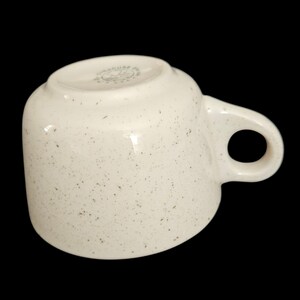 May include: Cream-colored ceramic mug with a speckled pattern and a rounded handle. The mug has a glossy finish and a slightly curved body. The bottom of the mug has the text "Syracuse China".