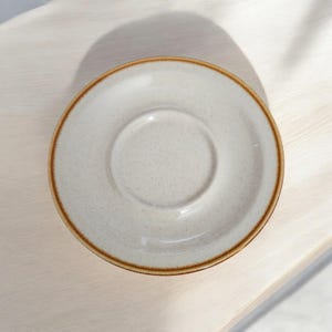 May include: A speckled, off-white ceramic saucer with a raised center and a brown rim. The saucer is round and sits on a light-colored wooden surface. The design is simple and elegant, suitable for a teacup or small cup.