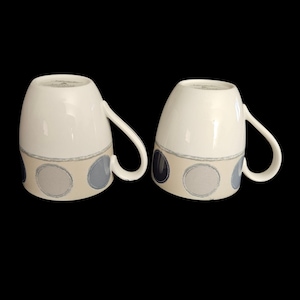 May include: Two white ceramic mugs with a beige band featuring gray and blue circles. Each mug has a curved handle and a slightly flared shape. The mugs are set against a black background.