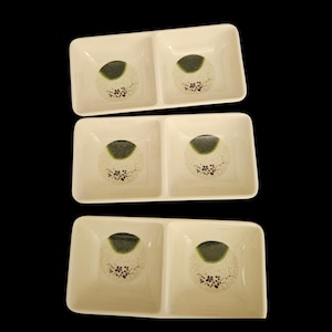 May include: Set of three off-white ceramic divided serving dishes. Each dish has two square compartments. The center of each compartment features a green and beige circular design with small floral accents. The dishes are arranged against a black background.