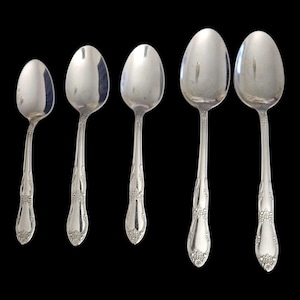 May include: Five silver spoons of varying sizes are arranged against a black background. The spoons have a reflective surface and ornate floral detailing on the handles. The spoons are likely for serving or eating.
