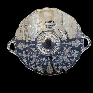 May include: Clear glass serving dish with a scalloped edge and etched floral design. The dish has two handles and a central raised section. The floral pattern includes roses and other flowers, creating an elegant, vintage aesthetic.