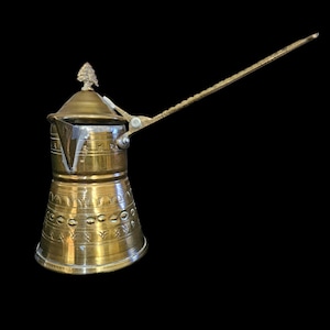 May include: A vintage brass coffee pot with an elongated handle and a decorative lid. The pot features etched patterns around the body and a small tree-like finial on the lid. The handle is long and textured.